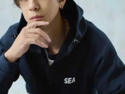 A Model wearing a navy zip-up hoodie with a small "SEA" logo, resting his chin on his hand in a close-up studio portrait