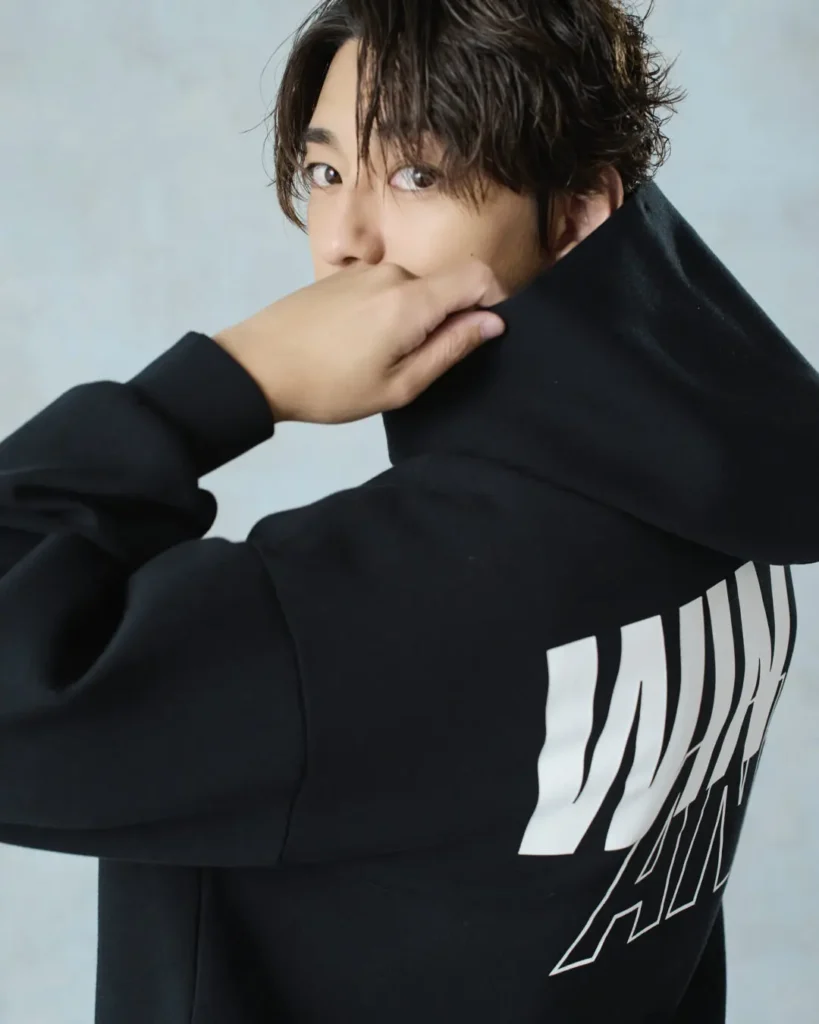 Close-up portrait of the model wearing a black hoodie with oversized "WIND AND SEA" branding across the back while glancing over his shoulder