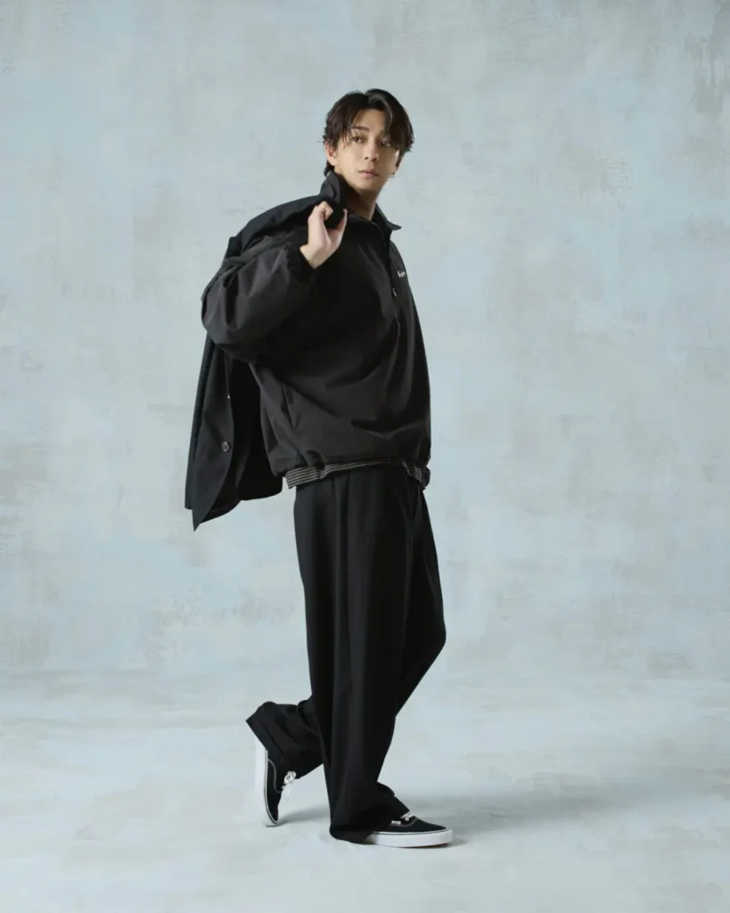 Full-body studio shot of the model in a black jacket, wide-leg trousers, and black sneakers with a jacket draped over one shoulder