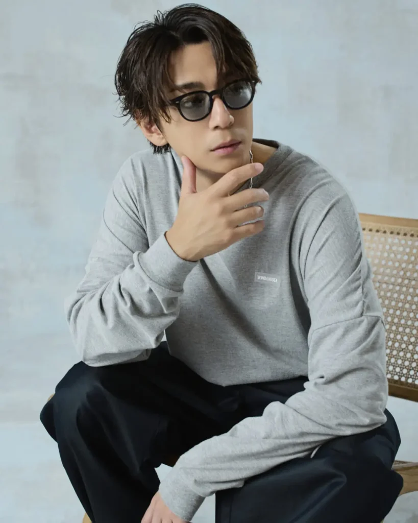 Seated portrait of the model wearing sunglasses, a gray "WIND AND SEA" sweatshirt, and black pants beside a woven chair