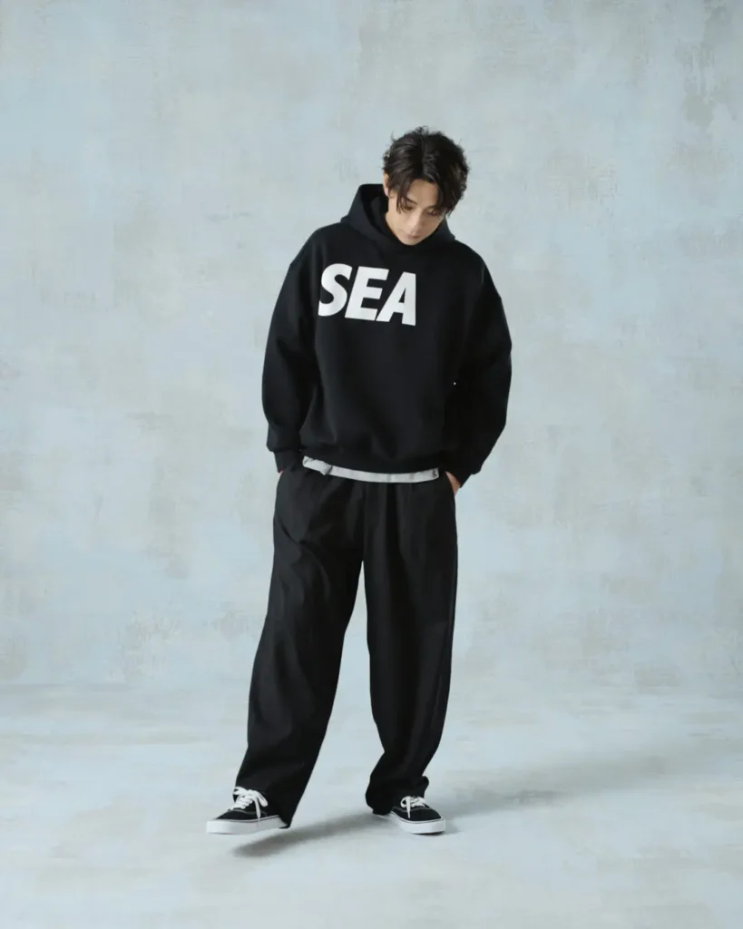 Full-body view of the model wearing a black "SEA" hoodie, relaxed black pants, and black canvas shoe