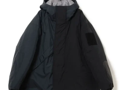 Black oversized hooded technical jacket with a high protective collar, concealed front closure, relaxed sleeves, and tonal utility patch