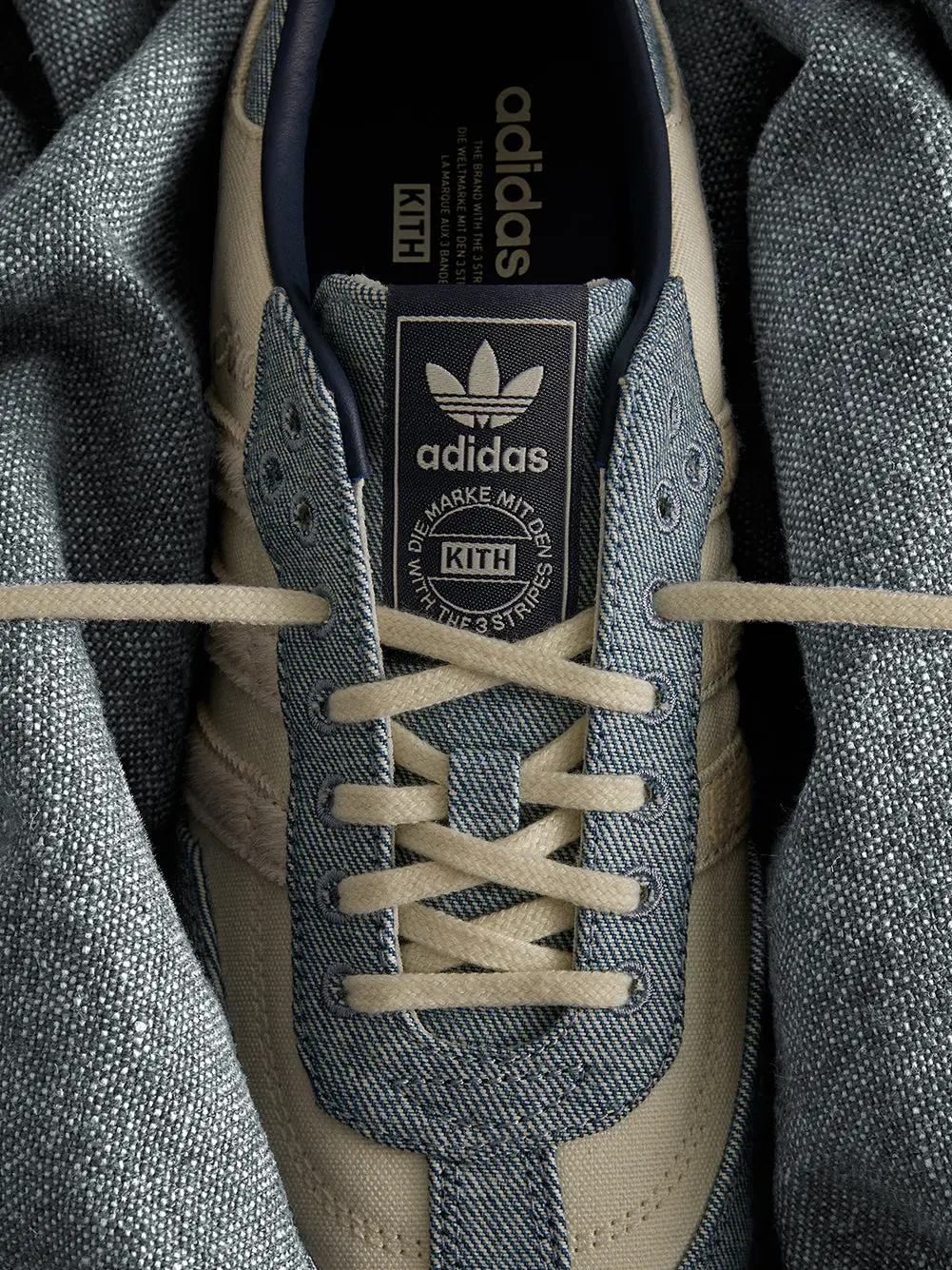 Close-up of a blue denim adidas sneaker with cream laces and KITH-branded tongue, framed by gray textured fabric