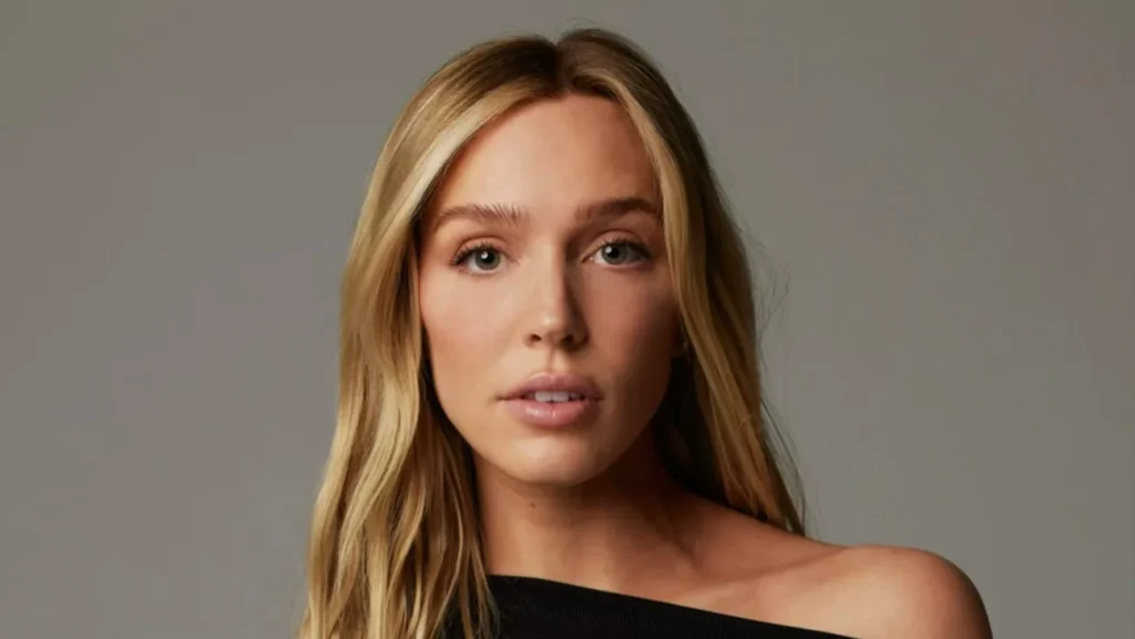 Close-up studio portrait of Alex Cooper with long, center-parted hair, wearing a black one-shoulder top and looking directly at the camera against a neutral gray background