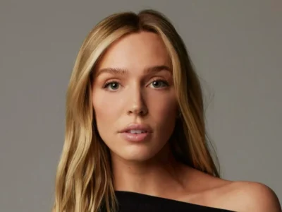 Close-up studio portrait of Alex Cooper with long, center-parted hair, wearing a black one-shoulder top and looking directly at the camera against a neutral gray background