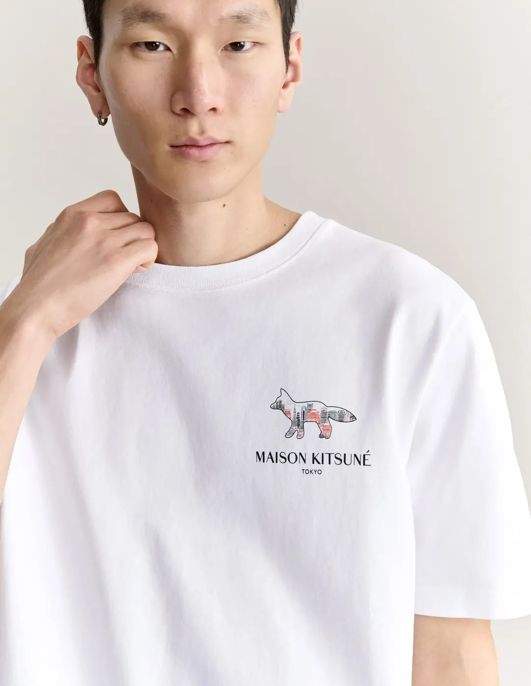 Model wearing a white Maison Kitsuné Tokyo T-shirt featuring a fox logo filled with a stylized Tokyo cityscape graphic on the left chest.