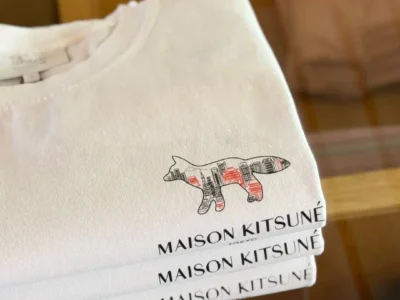 Folded white Maison Kitsuné Tokyo Tee stacked on a wooden chair, highlighting the embroidered fox cityscape logo and printed brand