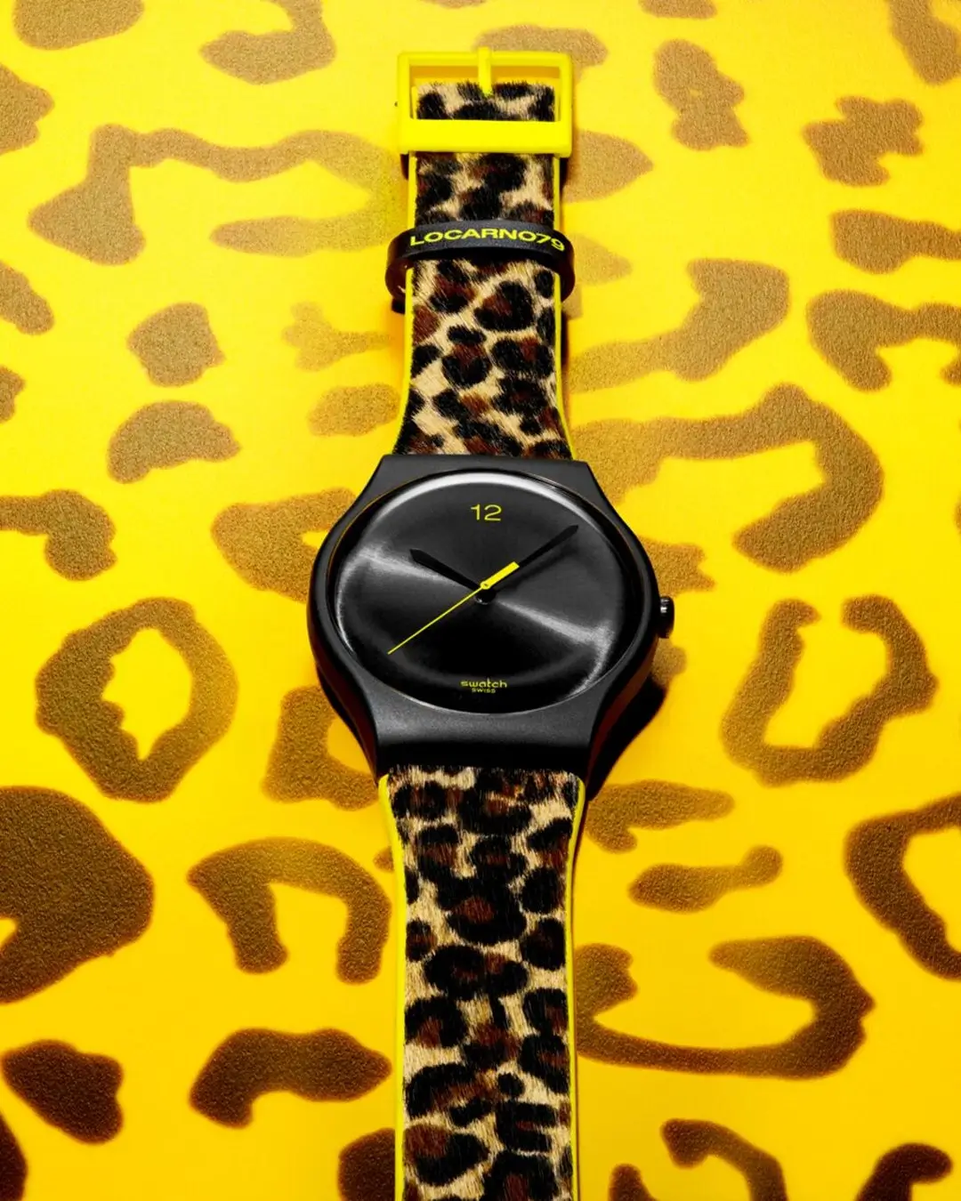 Front-facing Swatch watch with a matte black case, minimalist black dial, yellow hands, and a leopard-print hair-on strap displayed against a vibrant yellow leopard-pattern background.