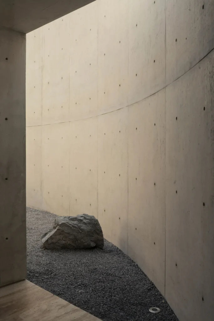 Min gravel courtyard framed by a gently curved exposed concrete wall, featuring a large natural boulder and soft daylight filtering from above