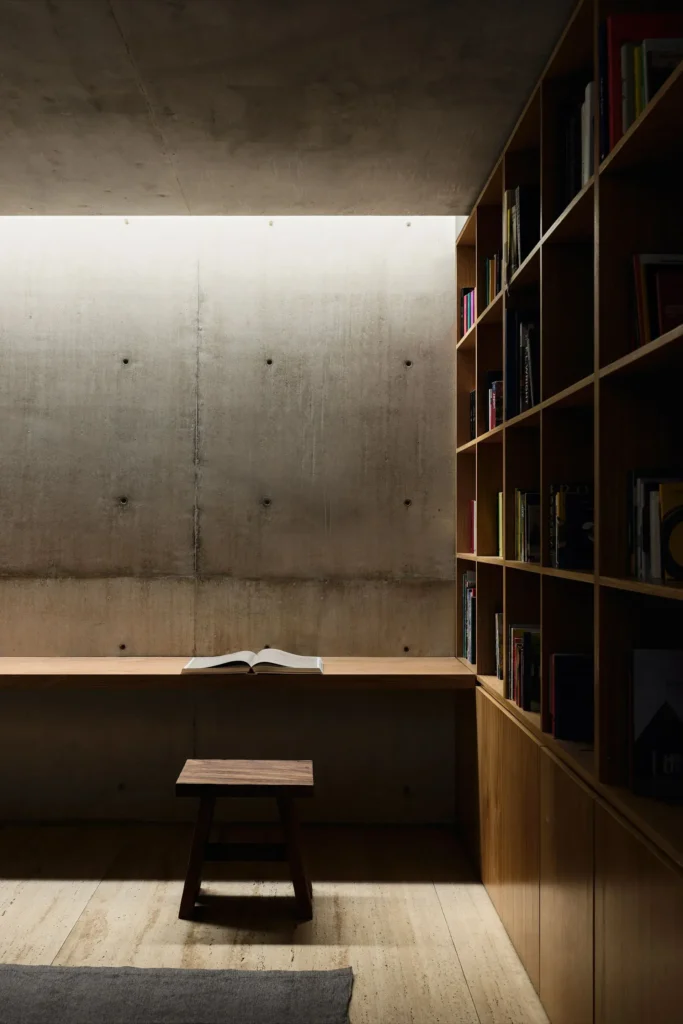 Quiet reading nook with floor-to-ceiling wooden bookshelves, an integrated desk, open book, and exposed concrete walls illuminated by natural skylight