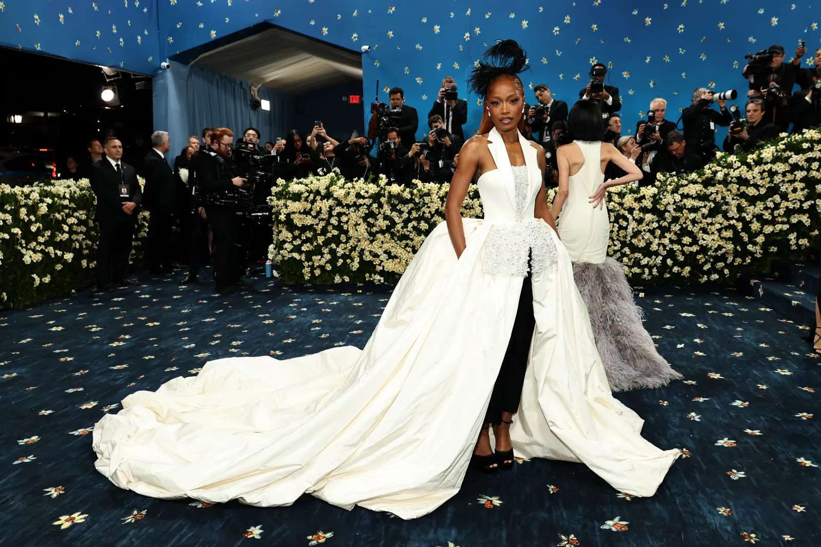 Woman posing on a floral blue carpet in a dramatic white gown with a jeweled structured bodice, voluminous overskirt, long train, black trousers, and sculptural feathered hair.