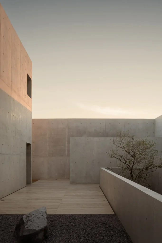 Minimalist rooftop courtyard with monolithic concrete volumes, warm stone paving, a sculptural tree, and soft golden-hour light beneath an open sky