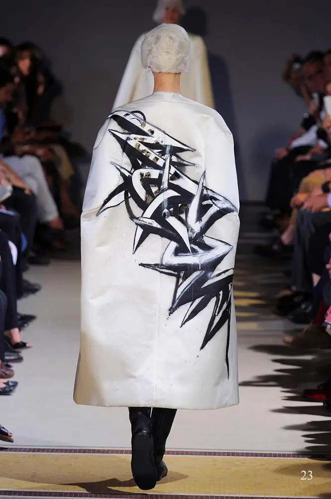 Rear runway view of a white Comme des Garçons coat covered with bold black abstract brushstroke graphics.