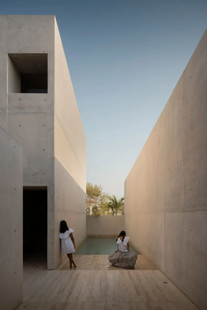 Narrow reflecting pool enclosed by tall exposed concrete walls, with two figures in white dresses interacting with the serene water and a natural stone