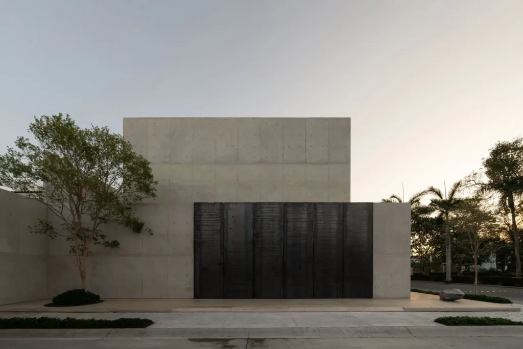 Front elevation of a minimal concrete residence featuring oversized black pivot doors, geometric architecture, mature trees, and a tranquil streetscape at dusk