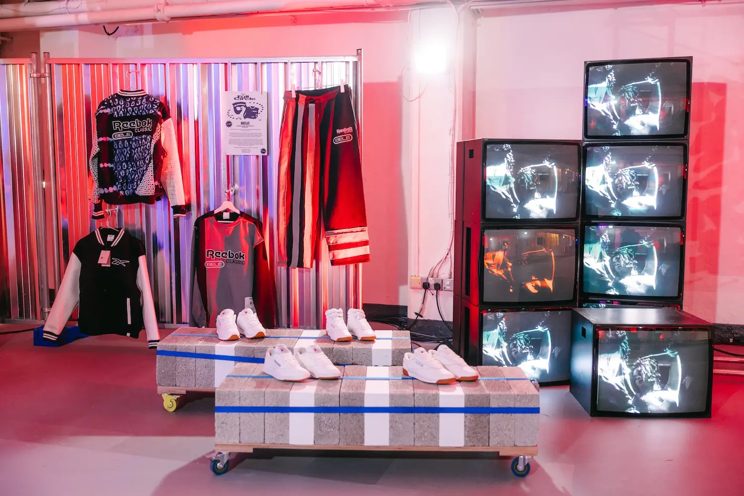 Reebok Classic apparel and footwear displayed in an industrial exhibition space, featuring graphic knitwear, track pants, varsity jackets, white sneakers on concrete plinths, and a wall of stacked CRT televisions playing campaign visuals under red ambient lighting.