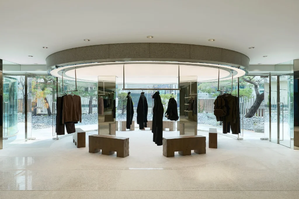 Circular Glass Fashion Display Area With Hanging Dark Garment And Stone Bench