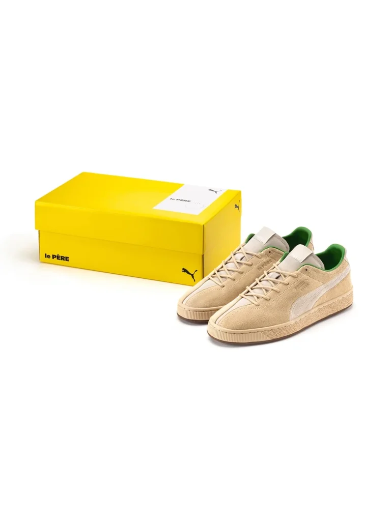 A product shot show a pair of PUMA x le PÈRE shoe displayed next to their collision box. The box is bright yellow with a black PUMA logo and a white label feature the "le PÈRE" brand. The shoe stand on their texture cream gum sole with bright green line and split-toe construction view in the foreground