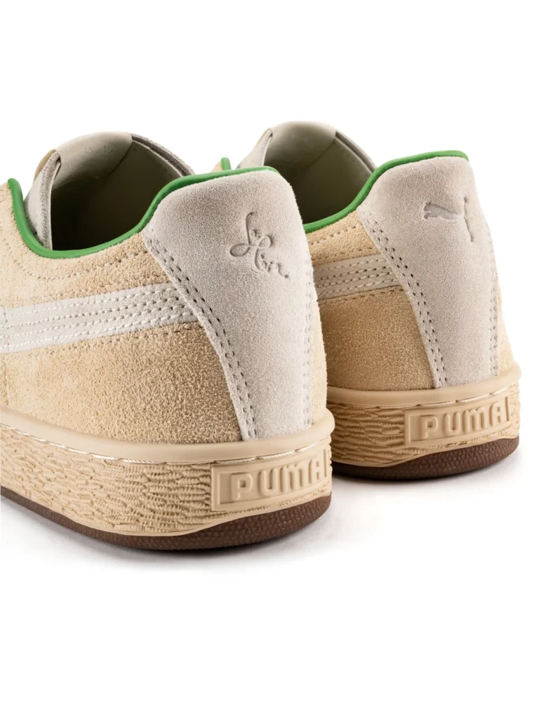 A close-up shot of the heel counter on the PUMA x le PÈRE Suede. A light silver-grey suede panel wraps around the back, feature an emboss PUMA logo on one shoe and a stylize le PÈRE logo on the other. The "PUMA" text is also emboss on the texture rear of the rubber sole, and the green line is view at the ankle