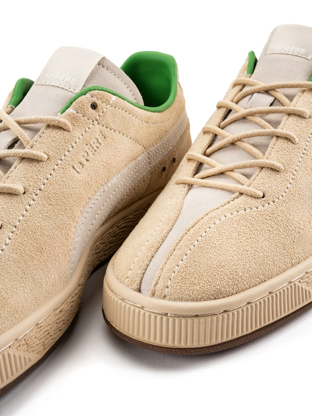 Close-up of two beige suede sneakers with green lining, tan laces, stitched panels, and ribbed rubber soles