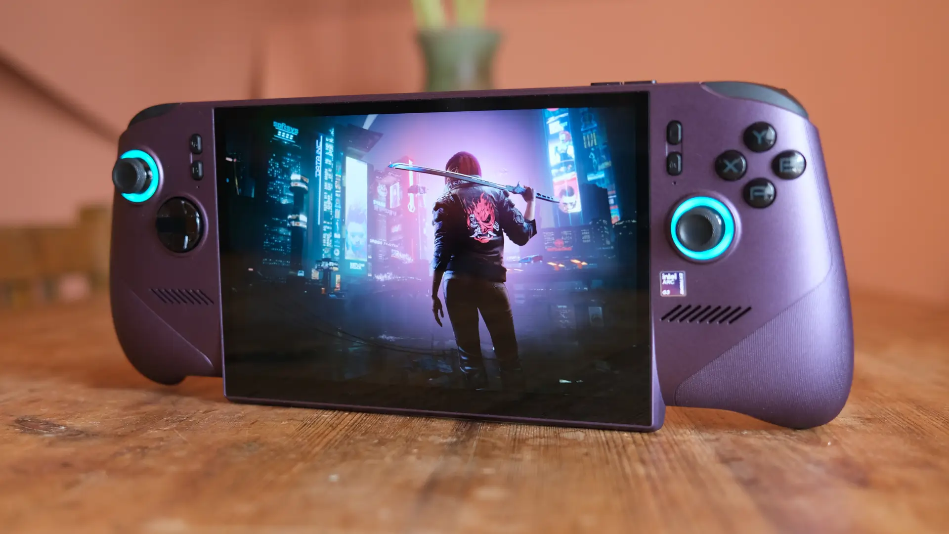 Purple handheld gaming console on a wooden tabletop displaying a neon-lit cyberpunk action scene, with glowing turquoise thumbsticks and a softly blurred indoor background.