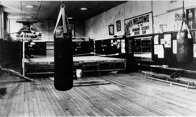 Black-and-white view of a historic boxing gym with a ring, heavy bags, wooden floors, and photographs lining the walls.