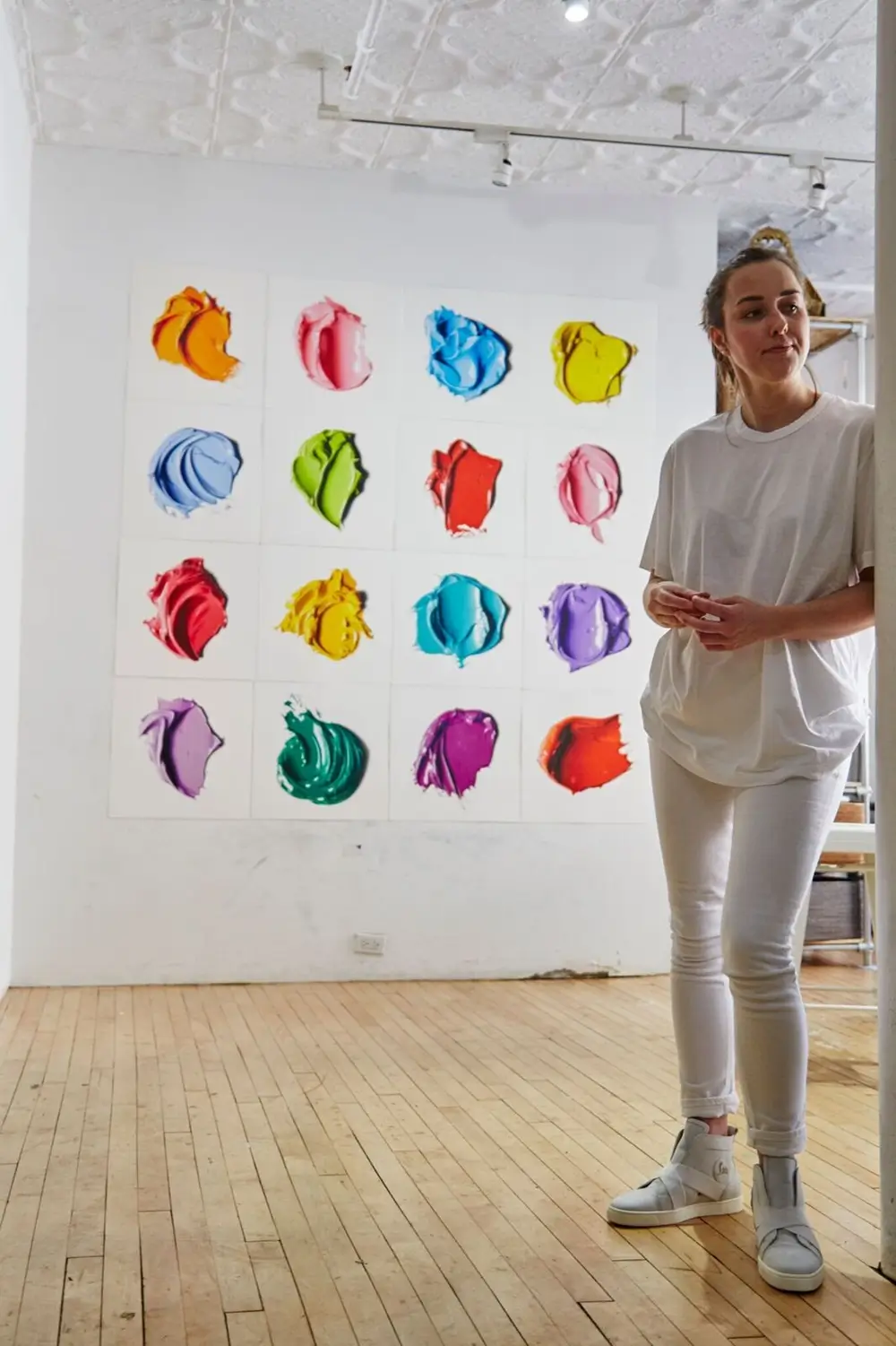 Artist Standing In A Bright White Studio Beside A Grid Of Sixteen Vivid Abstract Paintings Resembling Thick Swatches Of Colorful Paint.