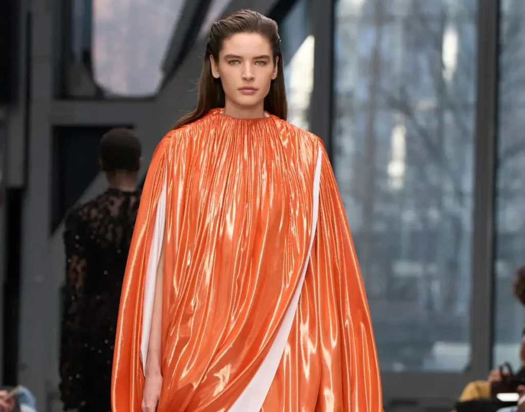 Model walking the runway in a glossy orange draped cape dress with sculptural volume and a high gathered neckline