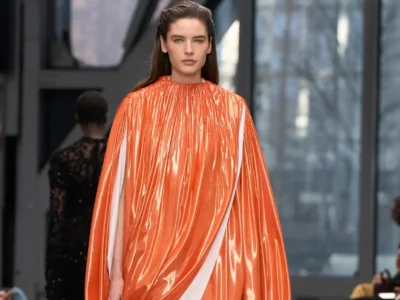 Model walking the runway in a glossy orange draped cape dress with sculptural volume and a high gathered neckline