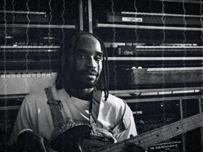 Monochrome portrait of a musician Dahi holding an electric bass in a studio lined with stacked synthesizers and keyboards, looking calmly toward the camera