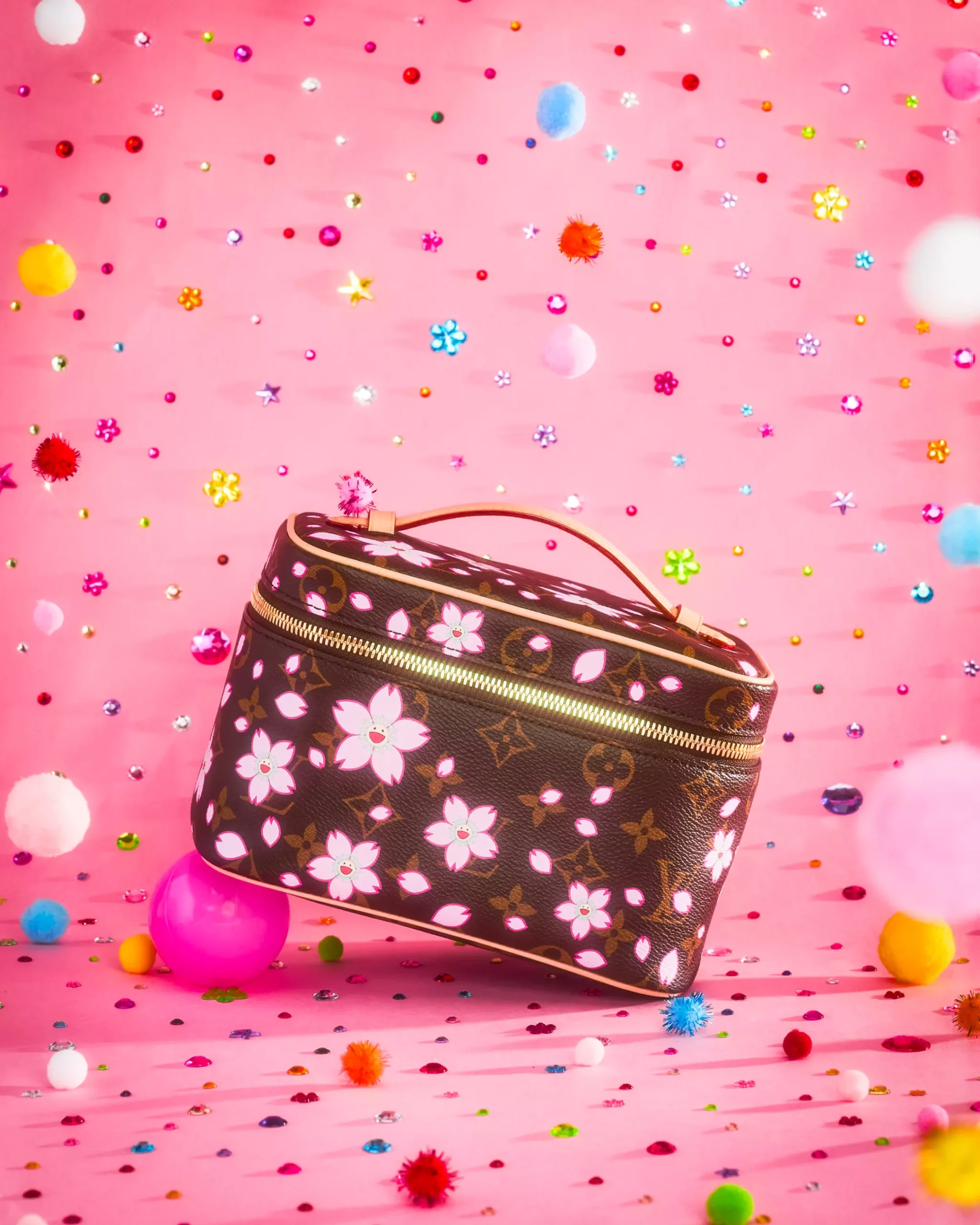 Louis Vuitton brown Monogram vanity bag decorated with pink cherry blossoms, natural leather trim, gold-tone zipper hardware, and a top handle, photographed against a bright pink background scattered with colorful gems and pom-poms.