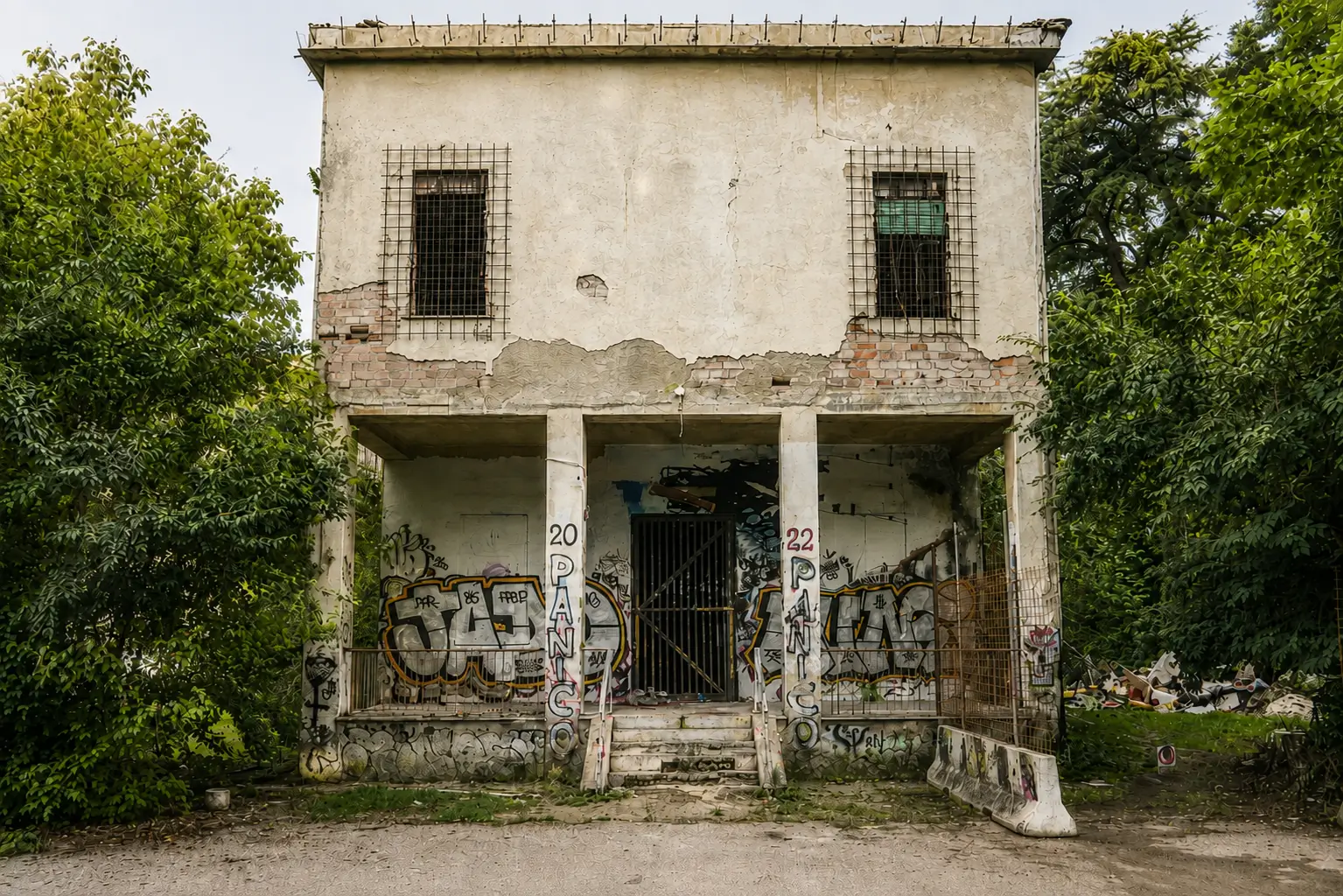 Front-facing view of a weathered abandoned two-story building with peeling plaster, exposed brick, graffiti-covered lower walls, barred windows, and dense trees framing the structure.