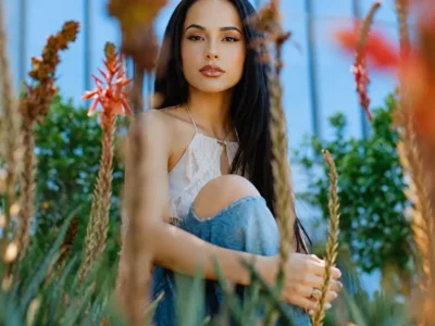 Becky G With Long Dark Hair Sit Among Lush Plants And Red Flowers, Wearing A White Halter Top And Blue Jeans