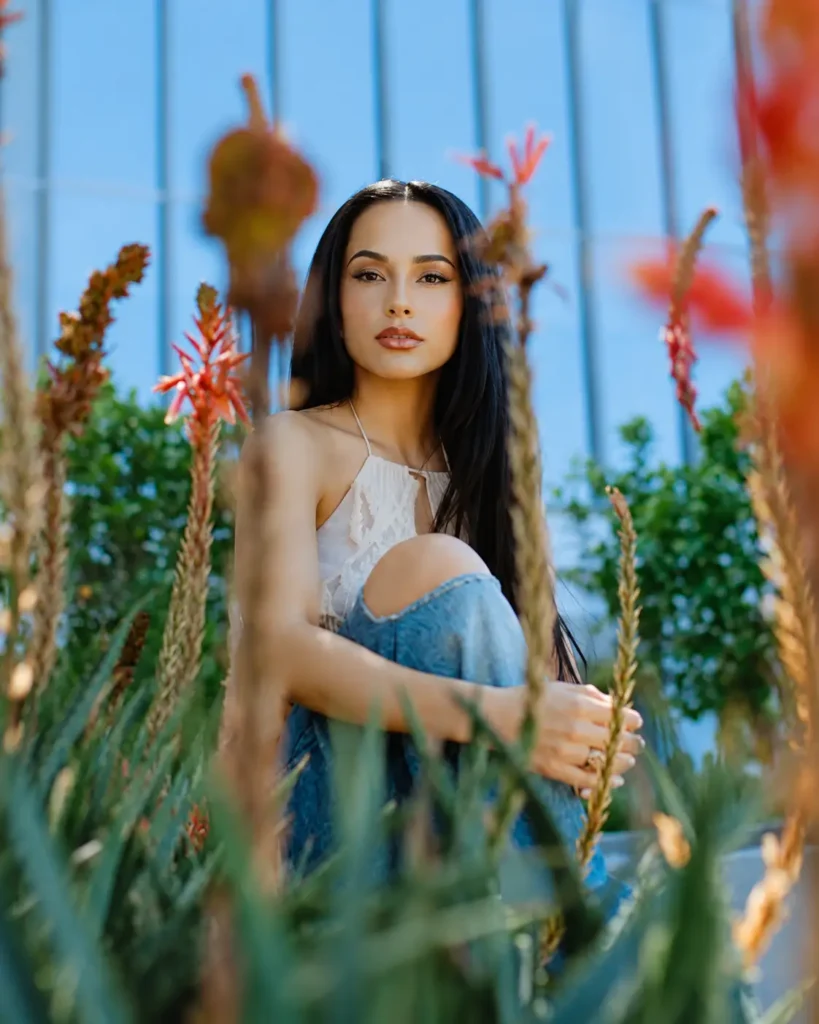 Becky G With Long Dark Hair Sit Among Lush Plants And Red Flowers, Wearing A White Halter Top And Blue Jeans