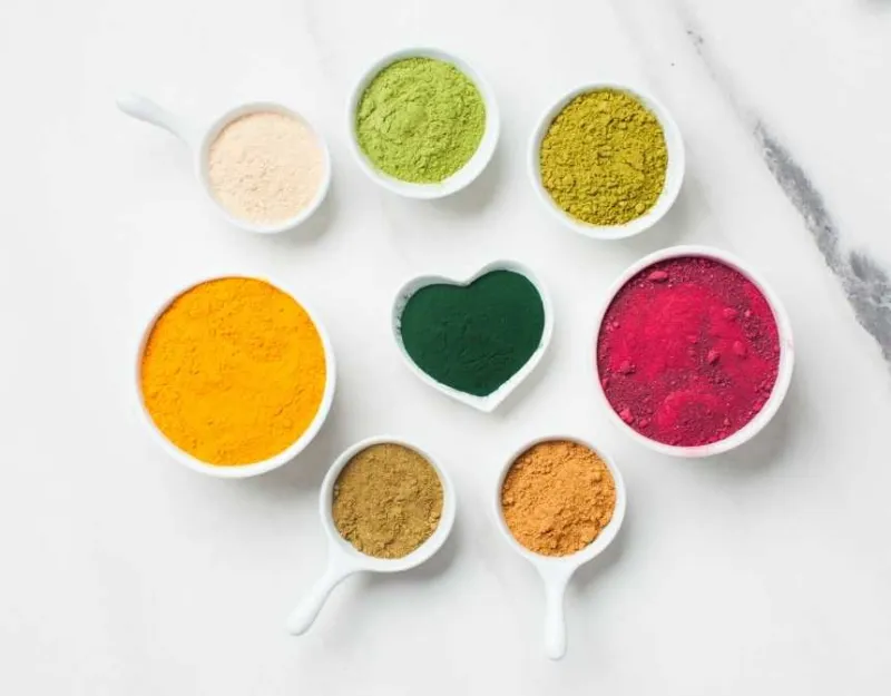 Bowls and spoons of brightly colored powdered ingredients arranged around a green heart-shaped dish on a white surface