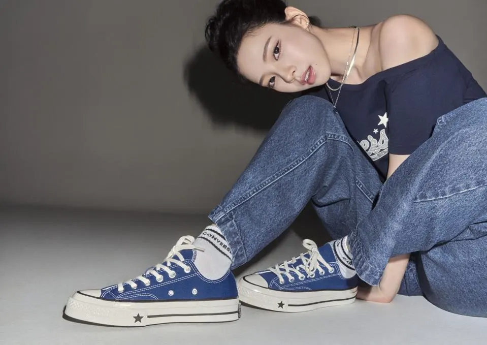 Model wearing a navy off-shoulder top, relaxed blue jeans, and blue Converse Chuck Taylor All Star sneakers in a minimalist studio portrait.