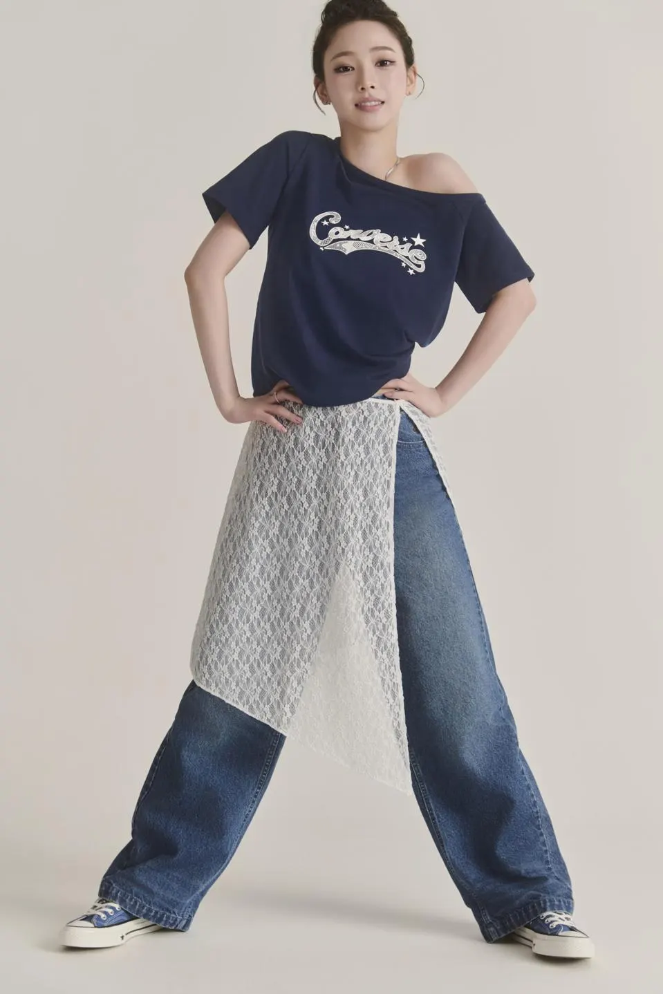 Person wears a navy Converse graphic T-shirt with wide-leg denim, a layered lace wrap skirt, and blue Converse Chuck Taylor sneakers in a minimalist studio.