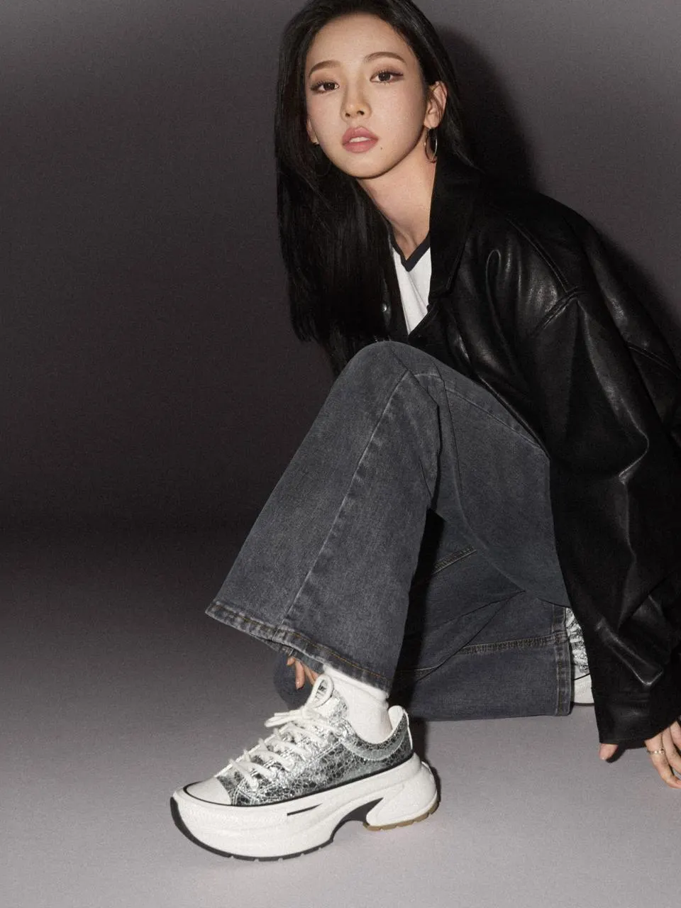 Person wearing a black leather jacket, white T-shirt, and gray flared jeans showcases silver textured Converse platform sneakers while seated against a dark studio backdrop.