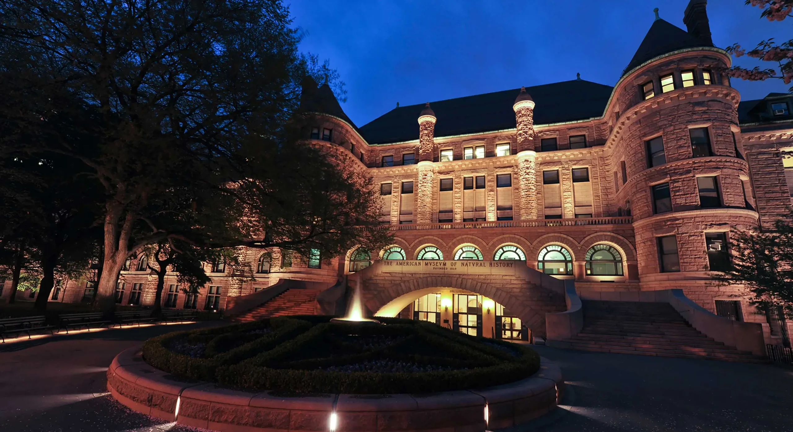 American Museum of Natural History illuminated at dusk, showing its historic stone facade, arched entrance, towers, landscaped courtyard, and surrounding trees.
