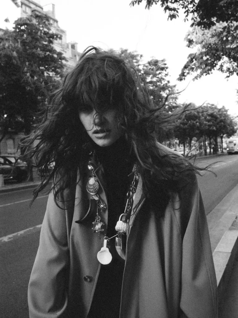 Long-haired model in a draped coat and layered necklaces stands on a tree-lined city street