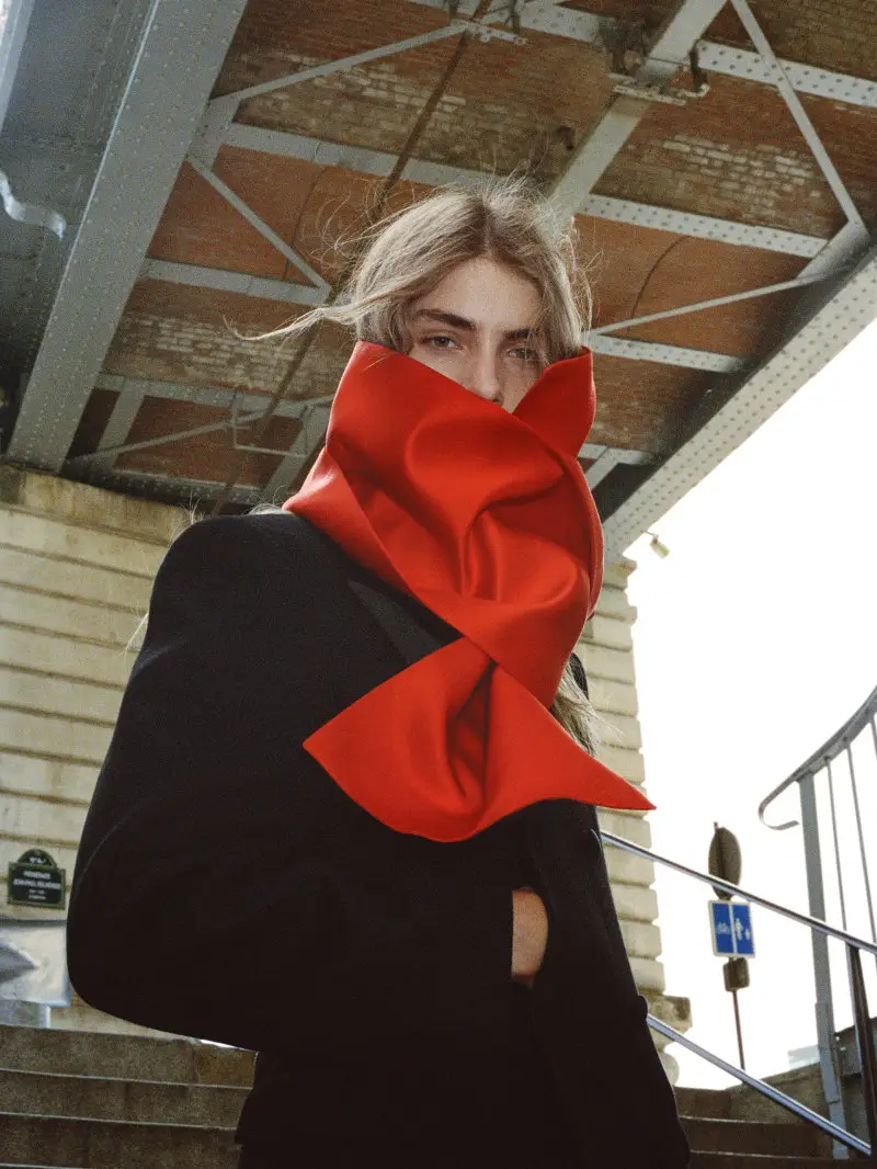 Person with tousled hair wears a black coat and oversized red scarf on an outdoor stairway beneath an industrial roof
