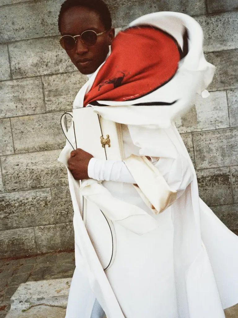 Model in white coat with red-lined hood, sunglasses and white shoulder bag against a stone wall