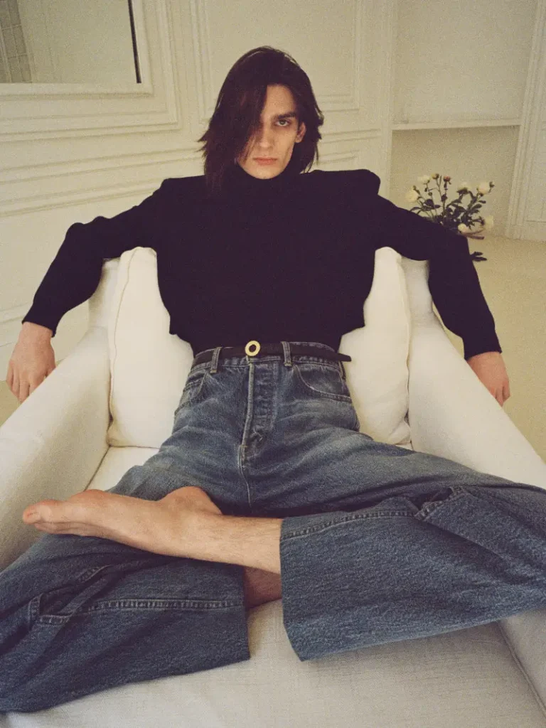 Long-haired man in a black top and wide-leg jeans sits barefoot in a white armchair, legs crossed and arms outstretched