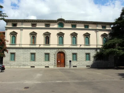 Classical masonry building with evenly spaced windows, ornate architectural detailing, and a broad paved forecourt in front