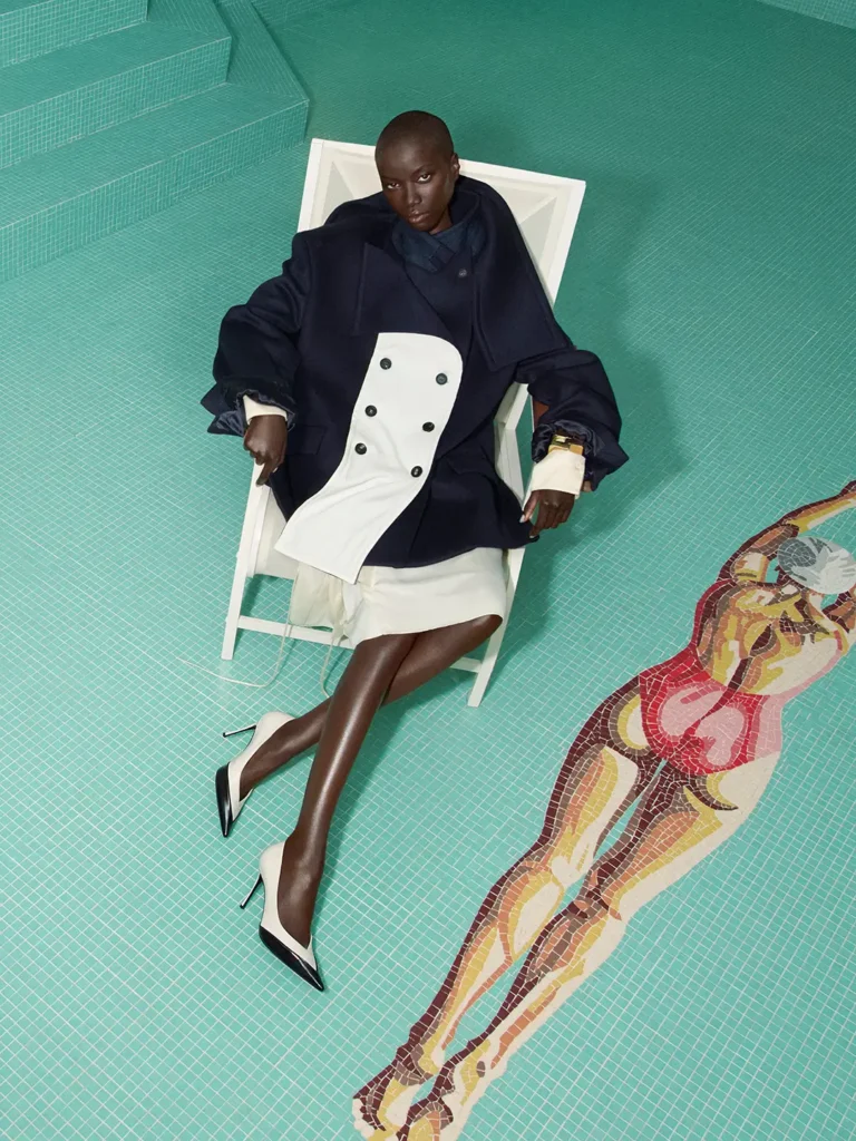 Model reclining beside a turquoise-tiled pool in an oversized navy-and-ivory coat, cream skirt, and black-and-white pointed pumps, creating a sculptural Ferragamo Fall/Winter editorial scene