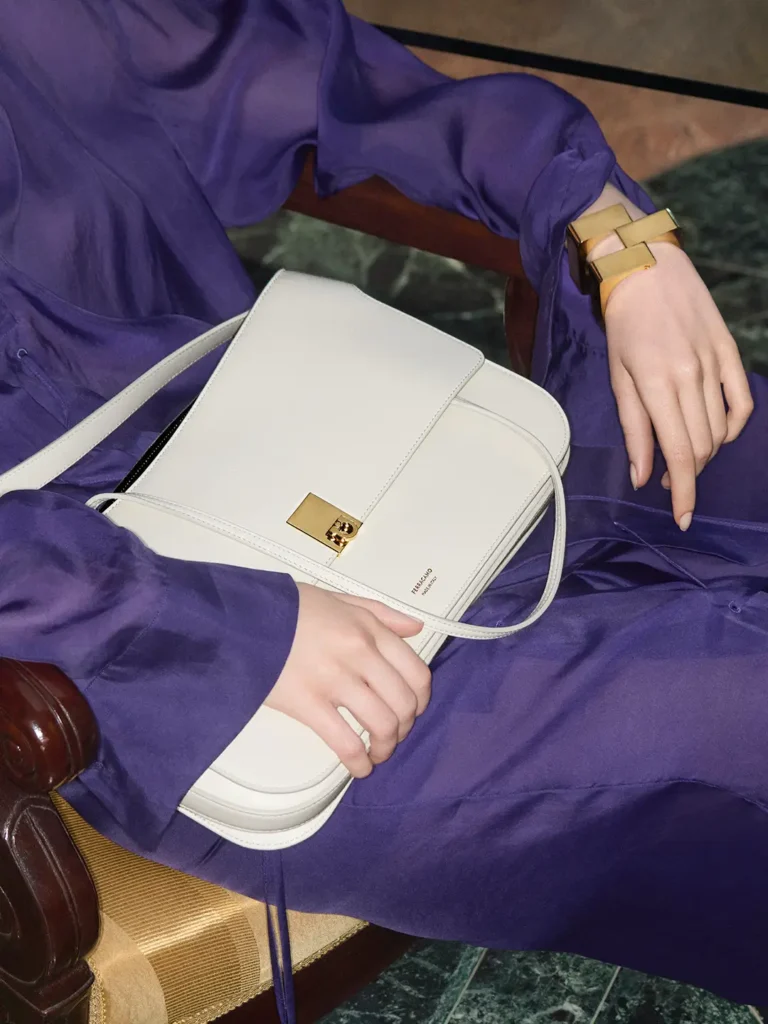Close-up of a structured white Ferragamo shoulder bag with gold-tone clasp resting across a violet dress, paired with a geometric gold cuff bracelet