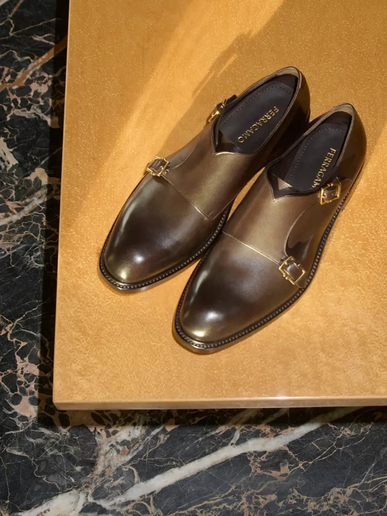 Pair of polished dark brown Ferragamo monk strap shoes with square gold buckles displayed on a warm gold tabletop with black marble surround
