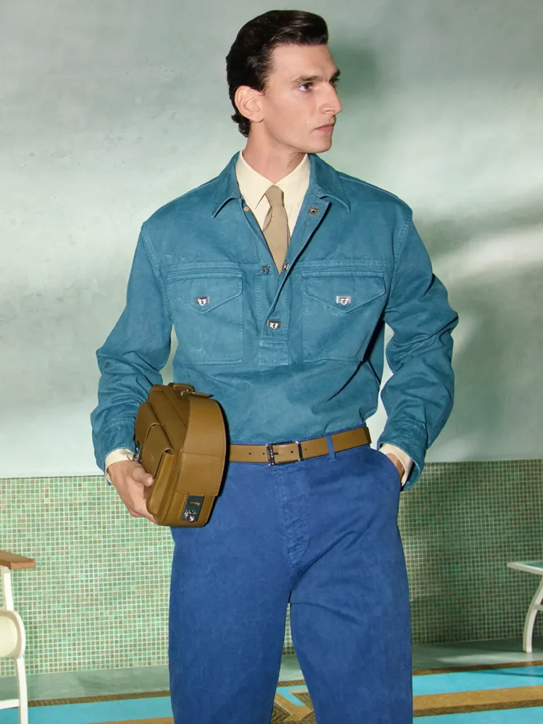 Male model wearing a teal overshirt, beige shirt and tie, cobalt trousers, and olive belt while carrying an olive Ferragamo crossbody bag in a min poolside set