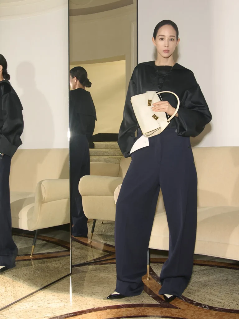 Model standing in wide-leg navy trousers and a black satin blouse, carrying a sculptural white Ferragamo handbag in a mirrored modern interior