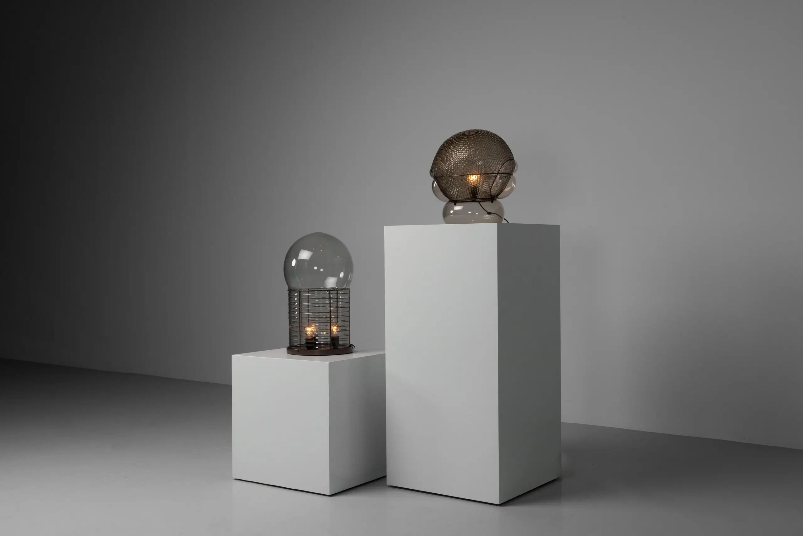Sculptural Glass And Metal Lamps Displayed On White Pedestals In A Minimal Gray Interior